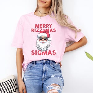 May include: A pink t-shirt with a graphic of Santa Claus wearing sunglasses and the text "Merry Rizzmas Sigmas".