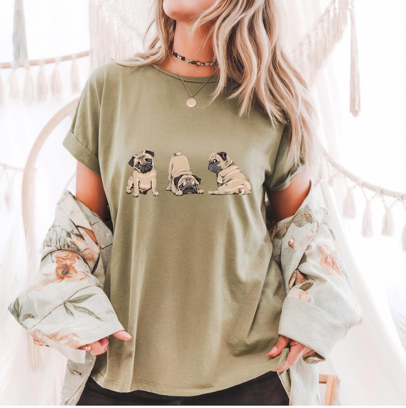 May include: Olive green t-shirt featuring a graphic of three pugs in different poses. The pugs are in various positions, including sitting, stretching, and lying down. The shirt has short sleeves and a relaxed fit.