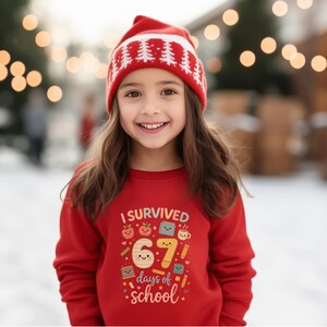 May include: A young person wearing a red sweatshirt and a red beanie with white tree designs. The sweatshirt has the text "I SURVIVED 67 days of school" with colorful graphics of apples, books, and pencils.