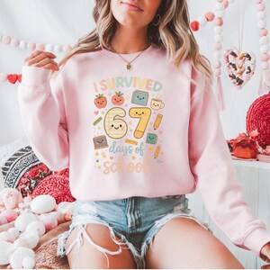 May include: A light pink sweatshirt with the text "I Survived 67 days of School" in a colorful, playful font. The graphic includes smiling cartoon images of a tomato, book, pencil, and coffee cup. The sweatshirt is a long-sleeved crew neck.