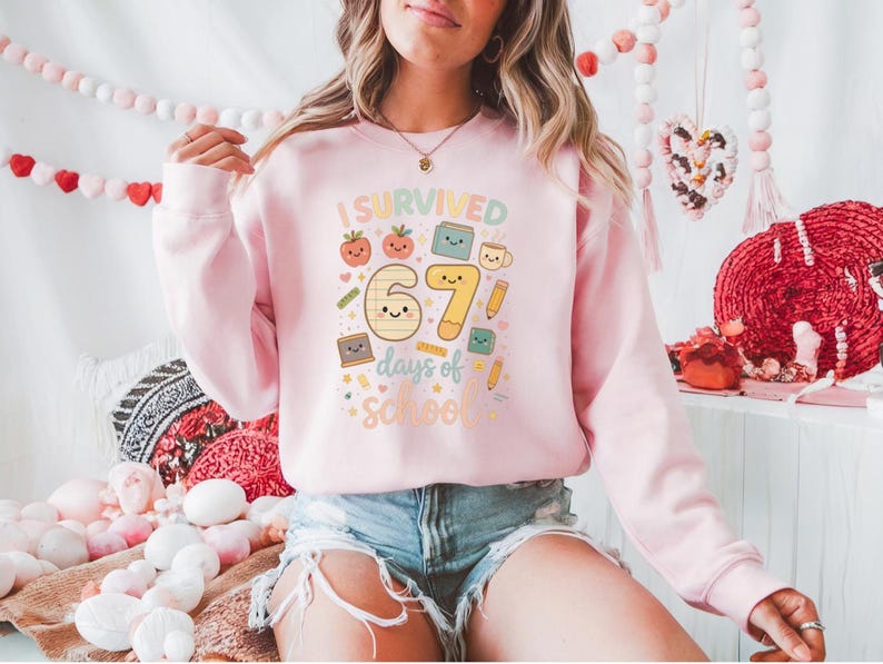 May include: A light pink sweatshirt with the text "I Survived 67 days of School" in a colorful, playful font. The graphic includes smiling cartoon images of a tomato, book, pencil, and coffee cup. The sweatshirt is a long-sleeved crew neck.