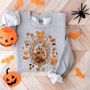 May include: A gray sweatshirt featuring a Halloween-themed design. The design includes a castle, pumpkins, Mickey Mouse ears, and fall-themed elements. Decorative orange bats and a jack-o'-lantern are placed around the sweatshirt.