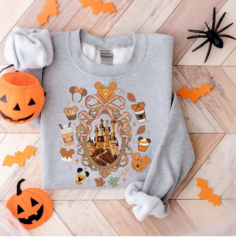 May include: A gray sweatshirt featuring a Halloween-themed design. The design includes a castle, pumpkins, Mickey Mouse ears, and fall-themed elements. Decorative orange bats and a jack-o'-lantern are placed around the sweatshirt.