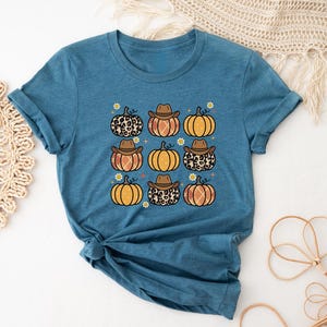 Puede incluir: Una camiseta azul turquesa con un gr&aacute;fico de nueve calabazas con sombreros de vaquero. Las calabazas son de color naranja, marr&oacute;n y tienen un estampado de leopardo.