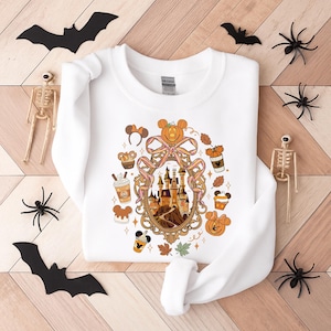 May include: White sweatshirt featuring a Halloween-themed design with a castle, pumpkins, and Mickey Mouse ears. The design is surrounded by fall leaves and coffee cups. Decorative bats, spiders, and skeletons are arranged around the sweatshirt.