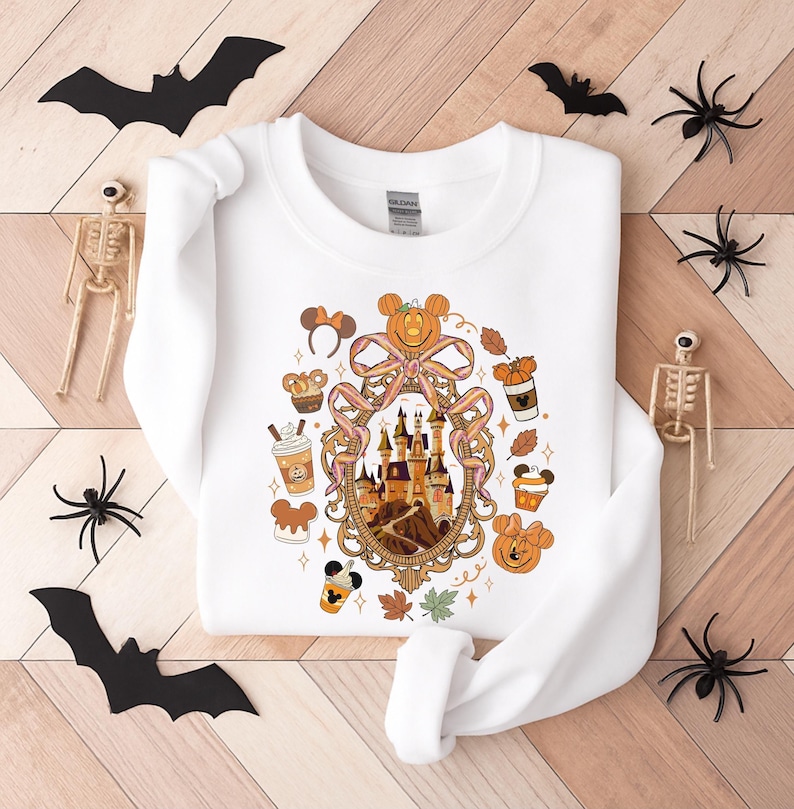 May include: White sweatshirt featuring a Halloween-themed design with a castle, pumpkins, and Mickey Mouse ears. The design is surrounded by fall leaves and coffee cups. Decorative bats, spiders, and skeletons are arranged around the sweatshirt.