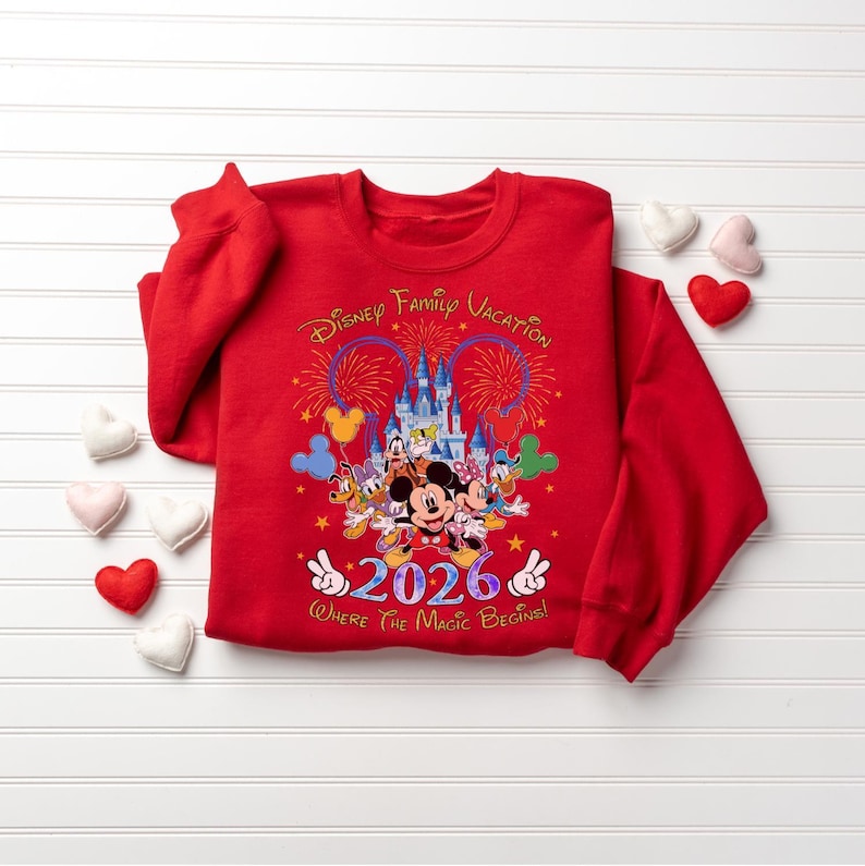 Peut inclure: Un sweat-shirt rouge avec un motif de vacances en famille Disney. Le graphisme comprend Mickey Mouse, Minnie Mouse, Donald Duck et d'autres personnages devant un ch&acirc;teau, avec l'ann&eacute;e 2026 et la phrase "Where The Magic Begins!"