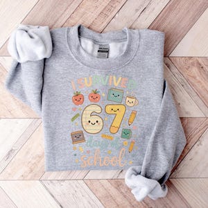 May include: A light gray sweatshirt with the text "I Survived 67 Days of School" in colorful letters and cute illustrations of apples, books, and pencils. The sweatshirt is made of a soft material and is perfect for students.