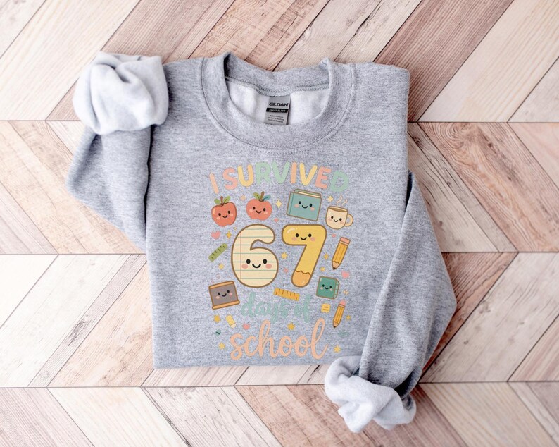 May include: A light gray sweatshirt with the text "I Survived 67 Days of School" in colorful letters and cute illustrations of apples, books, and pencils. The sweatshirt is made of a soft material and is perfect for students.