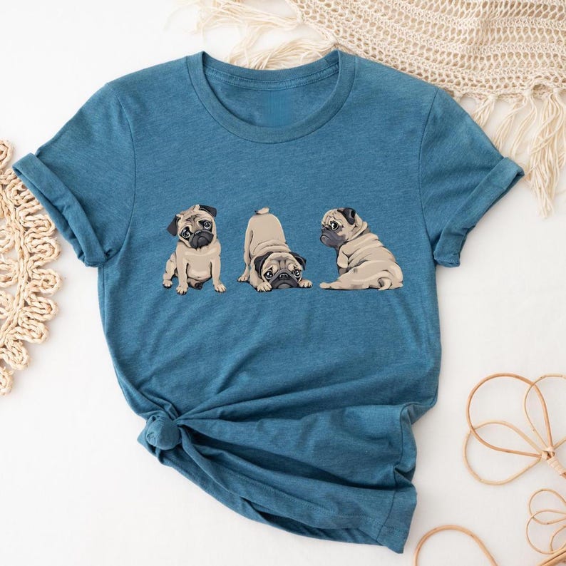 May include: A blue t-shirt featuring three cartoon pugs in different poses. The pugs are tan with black faces and ears. The shirt has short sleeves and a round neckline. The fabric appears to be a soft, lightweight material.