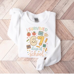 May include: White sweatshirt with the text "I Survived 67 days of school" in colorful, playful lettering. The design includes cartoon illustrations of apples, books, pencils, and a coffee cup, perfect for back-to-school or end-of-year celebrations.