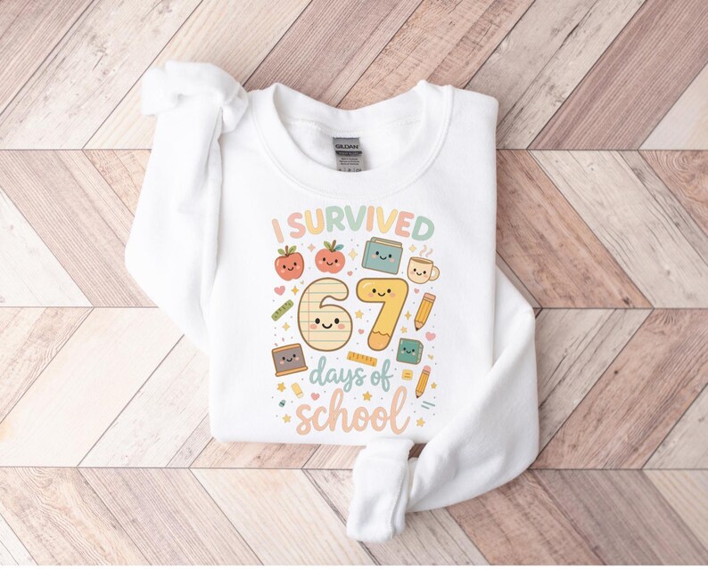 May include: White sweatshirt with the text "I Survived 67 days of school" in colorful, playful lettering. The design includes cartoon illustrations of apples, books, pencils, and a coffee cup, perfect for back-to-school or end-of-year celebrations.