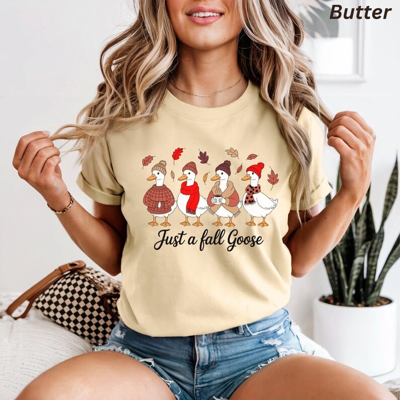 May include: Butter-colored t-shirt featuring a graphic of four geese dressed in fall attire, including hats, scarves, and sweaters. The text "Just a fall Goose" is printed below the geese. The shirt has short sleeves.