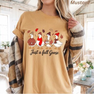 May include: Mustard-colored t-shirt featuring a graphic of four geese dressed in fall attire, including hats, scarves, and sweaters. The text "Just a fall Goose" is printed below the geese. The shirt has rolled-up sleeves.