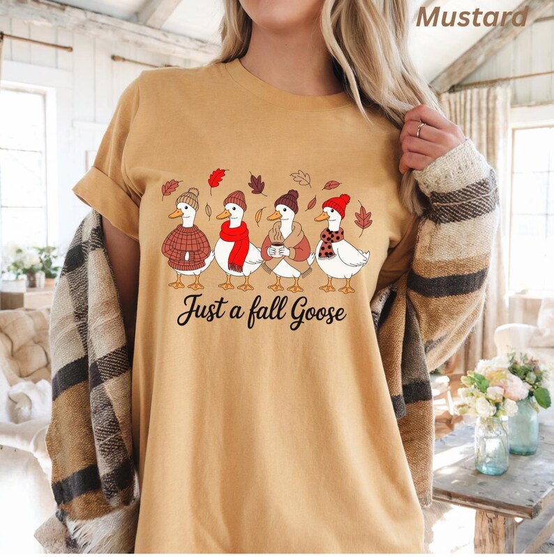 May include: Mustard-colored t-shirt featuring a graphic of four geese dressed in fall attire, including hats, scarves, and sweaters. The text "Just a fall Goose" is printed below the geese. The shirt has rolled-up sleeves.