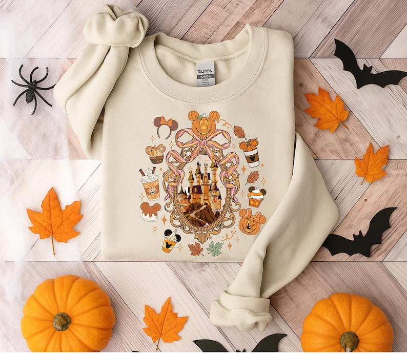 May include: Cream-colored sweatshirt featuring a framed illustration of a castle, pumpkins, and fall-themed elements. The design includes Mickey Mouse ears, coffee cups, and autumn leaves. The sweatshirt is surrounded by decorative pumpkins, leaves, and bats.