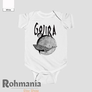 May include: White baby bodysuit with a black graphic. The design features the word "GOJIRA" above a detailed illustration of a whale swimming in front of a textured sphere. The bodysuit is a one-piece garment.