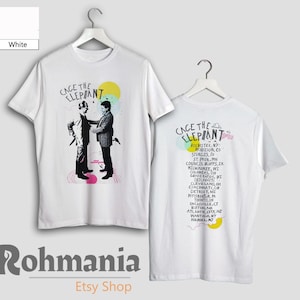 May include: White t-shirts with a graphic of two figures and the text "CAGE THE ELEPHANT". The back of the shirt features the band's name and a list of tour locations. The shirts are displayed on hangers.