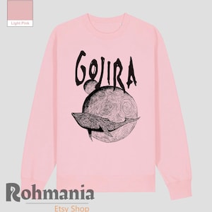May include: Light pink sweatshirt with a black graphic. The graphic features the word "GOJIRA" above a detailed illustration of a whale swimming in front of a planet-like design. The Etsy shop name "Rohmania" is at the bottom.