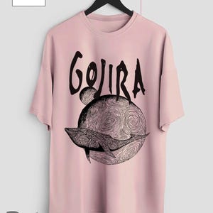 Op de afbeelding: Lichtroze t-shirt met het woord "GOJIRA" in zwart boven een zwart-wit illustratie van een walvis op een wervelende planeet. Het t-shirt hangt aan een zwarte hanger.