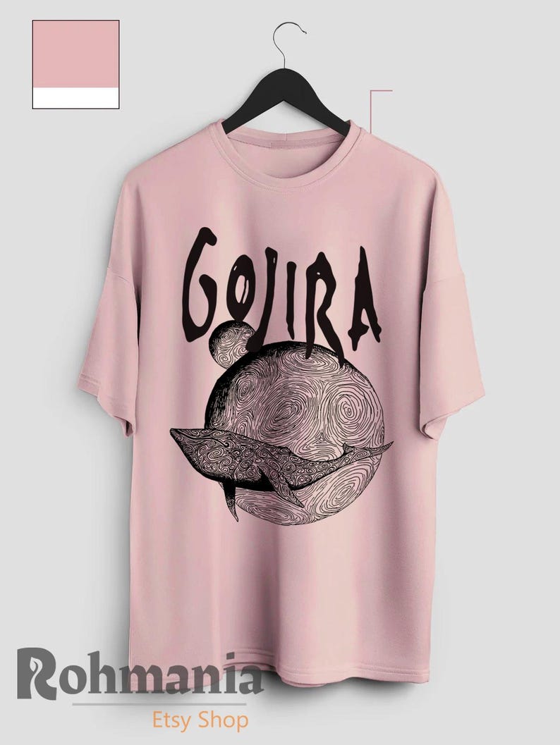 May include: A light pink t-shirt featuring the word "GOJIRA" in a black, stylized font above a black and white illustration of a whale on a swirling planet. The shirt is hanging on a black hanger.