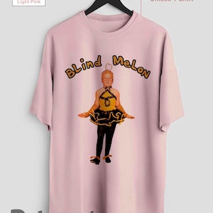 Puede incluir: Camiseta unisex rosa claro con la inscripción "Blind Melon" en amarillo y negro. La camiseta tiene un gráfico de una persona con un atuendo amarillo y negro. La camiseta está colgada en una percha negra.