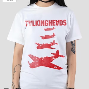 Puede incluir: Camiseta blanca con el nombre de la banda "TALKINGHEADS" en rojo sobre una serie de gráficos de aviones rojos. La camiseta es de cuello redondo y manga corta.