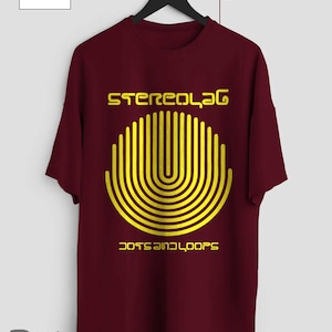 Puede incluir: Una camiseta granate con un diseño gráfico amarillo. El diseño presenta el texto "Stereolab" sobre un patrón semicircular de líneas verticales y el texto "Dots and Loops" debajo. La camiseta está colgada de una percha negra.