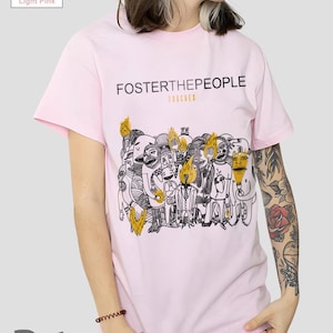 Puede incluir: Camiseta rosa claro con el nombre de la banda "Foster the People" y la palabra "Torches" sobre un gráfico negro y amarillo de figuras estilizadas. La camiseta es de color rosa claro.