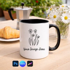 May include: A white ceramic mug with a black handle and interior. The mug features a floral design and the text "Your Design Here". The image also includes a croissant, a coffee maker, and a vase of daisies.