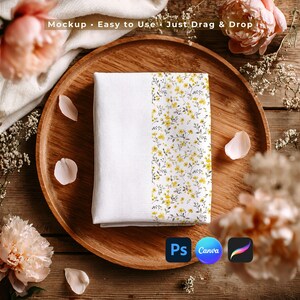 Puede incluir: Un paño blanco doblado con un estampado floral de flores amarillas y hojas verdes, colocado sobre una bandeja de madera. El texto "Mockup • Easy to Use • Just Drag & Drop" es visible. La imagen también incluye los logotipos de Photoshop, Canva y Procreate.