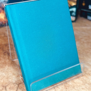 May include: A teal blue hardcover book on a clear acrylic book stand. The book is on a light brown surface.