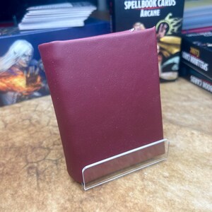 May include: A burgundy leather card holder with a clear acrylic stand. The card holder is sitting on a brown surface. The text "D&D Spellbook Cards Arcane" is visible on a box in the background.