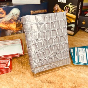 May include: A white card deck box with a crocodile skin pattern. The box is made of a sturdy material and has a clear plastic stand.