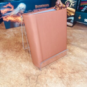 May include: A brown leather-bound book with a clear acrylic stand. The book is closed and has a smooth surface. The stand is clear and has a rectangular shape.