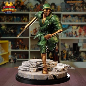 May include: A detailed figurine of a soldier in a dynamic pose, clad in a green uniform with gold-colored mechanical limbs and helmet. The figure is holding a rifle and stands on a rocky base. The logo "LEGION MINIATURES 3D PRINTING DESIGNS" is visible.