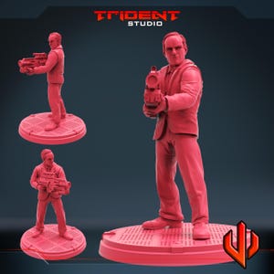 Może przedstawiać: Czerwona figurka 3D przedstawiająca mężczyznę w garniturze trzymającego futurystyczną broń. Figurka stoi na okrągłej podstawie o teksturowanej powierzchni. Widoczne jest logo Trident Studio.