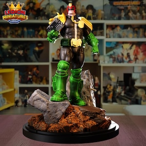 May include: A detailed figurine of Judge Dredd, a comic book character, standing on a rocky base. The figure is predominantly black, green, and gold, with a helmet and a futuristic weapon. The base is dark brown and the figure is on a black circular base.