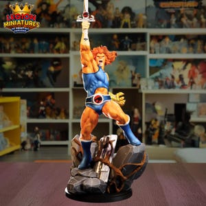 May include: A detailed figurine of a muscular, orange-haired character in a blue bodysuit, raising a sword. The figure stands on a rocky base with a logo that reads "LEGION MINIATURES 3D PRINTING DESIGNS".