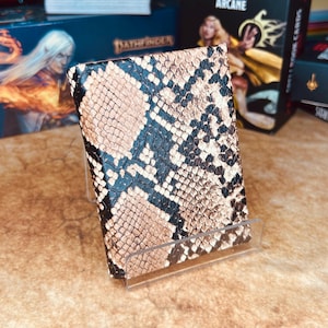 May include: A brown and black snakeskin-patterned card case with a clear acrylic stand. The case is made of faux leather and has a textured surface.