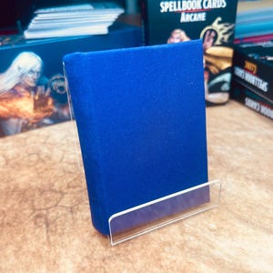 May include: A blue, cloth-bound book on a clear acrylic stand. The book is titled "D&D Spellbook Cards Arcane".