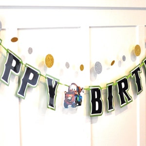 May include: A festive "Happy Birthday" banner with black letters outlined in green. The banner includes a cartoon image of a car and is strung with a green cord. Gold and silver glitter confetti dots are scattered around the banner.