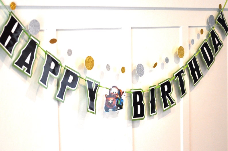 May include: A festive "Happy Birthday" banner with black letters outlined in green. The banner includes a cartoon image of a car and is strung with a green cord. Gold and silver glitter confetti dots are scattered around the banner.