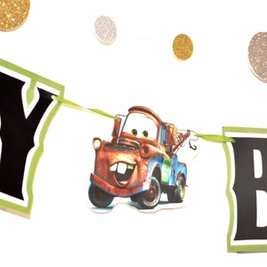 May include: A banner with the word "Y B" in black letters with a green border, featuring a cartoon image of Tow Mater from the movie Cars. The banner is strung with green ribbon and has gold and silver glitter circle decorations.