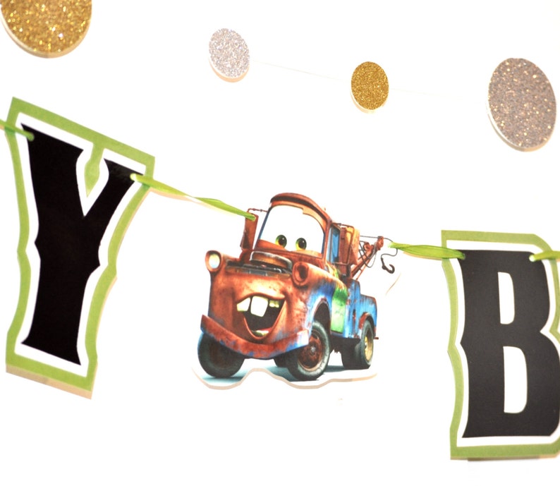 May include: A banner with the word "Y B" in black letters with a green border, featuring a cartoon image of Tow Mater from the movie Cars. The banner is strung with green ribbon and has gold and silver glitter circle decorations.