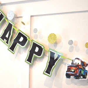 May include: A festive banner spelling "HAPPY" in black letters with a green border, strung on a green ribbon. The banner includes a cartoon image of a tow truck. Gold and silver glitter circles are also strung on the ribbon.