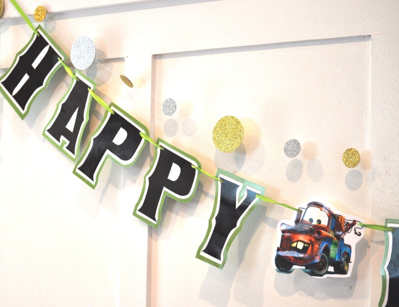 May include: A festive banner spelling "HAPPY" in black letters with a green border, strung on a green ribbon. The banner includes a cartoon image of a tow truck. Gold and silver glitter circles are also strung on the ribbon.