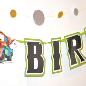 May include: A birthday banner with the word "BIRTH" in black letters with a green border. The banner includes a cartoon image of a red and blue tow truck. Glittery gold and silver circles are strung above the banner.