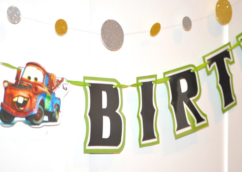 May include: A birthday banner with the word "BIRTH" in black letters with a green border. The banner includes a cartoon image of a red and blue tow truck. Glittery gold and silver circles are strung above the banner.