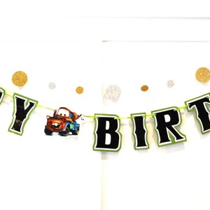 May include: A festive "HAPPY BIRTHDAY" banner with black letters outlined in white and green, strung on a green cord. The banner includes a cartoon image of a red and blue truck. Gold and silver confetti dots are scattered around the banner.